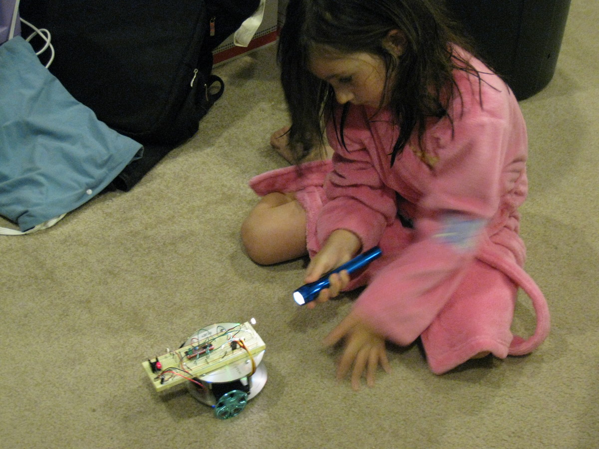 Young Sarah Orner sitting next to Roby the Costerbot, a small Arduino robot built on a CD platter platform, demonstrating early STEM learning in 2010